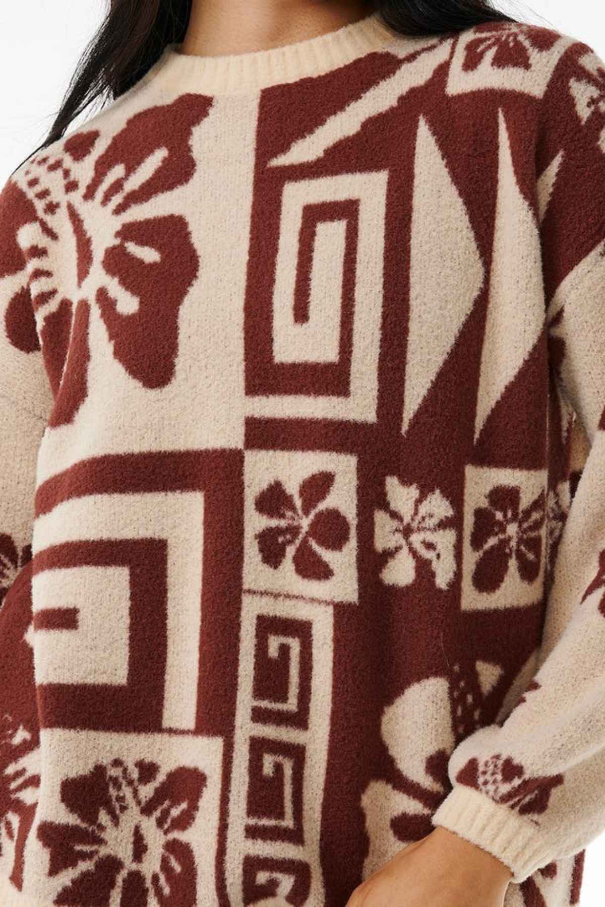 A person is dressed in the comfortable Rip Curl Block Party Jacquard Crew sweater, featuring a unique abstract floral and geometric pattern in shades of burgundy and cream. The jacquard knit design showcases large flowers, spirals, and various shapes. The person's face remains unseen, highlighting the sweater's detailed craftsmanship.