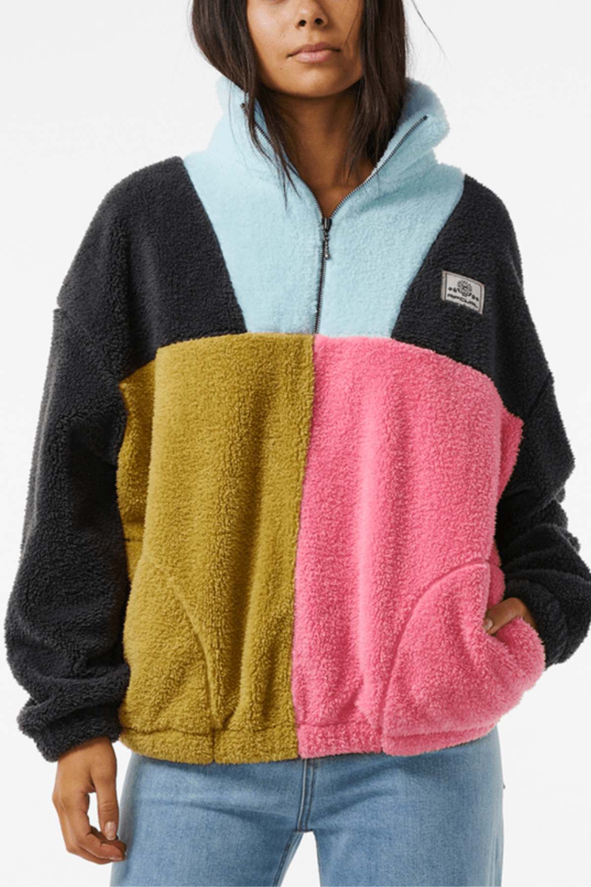 A person wears a Rip Curl Block Party Polar Fleece with a quarter-zip in black, light blue, olive green, and pink color blocks. A patch adorns the chest, making it an ideal pairing with light blue jeans.
