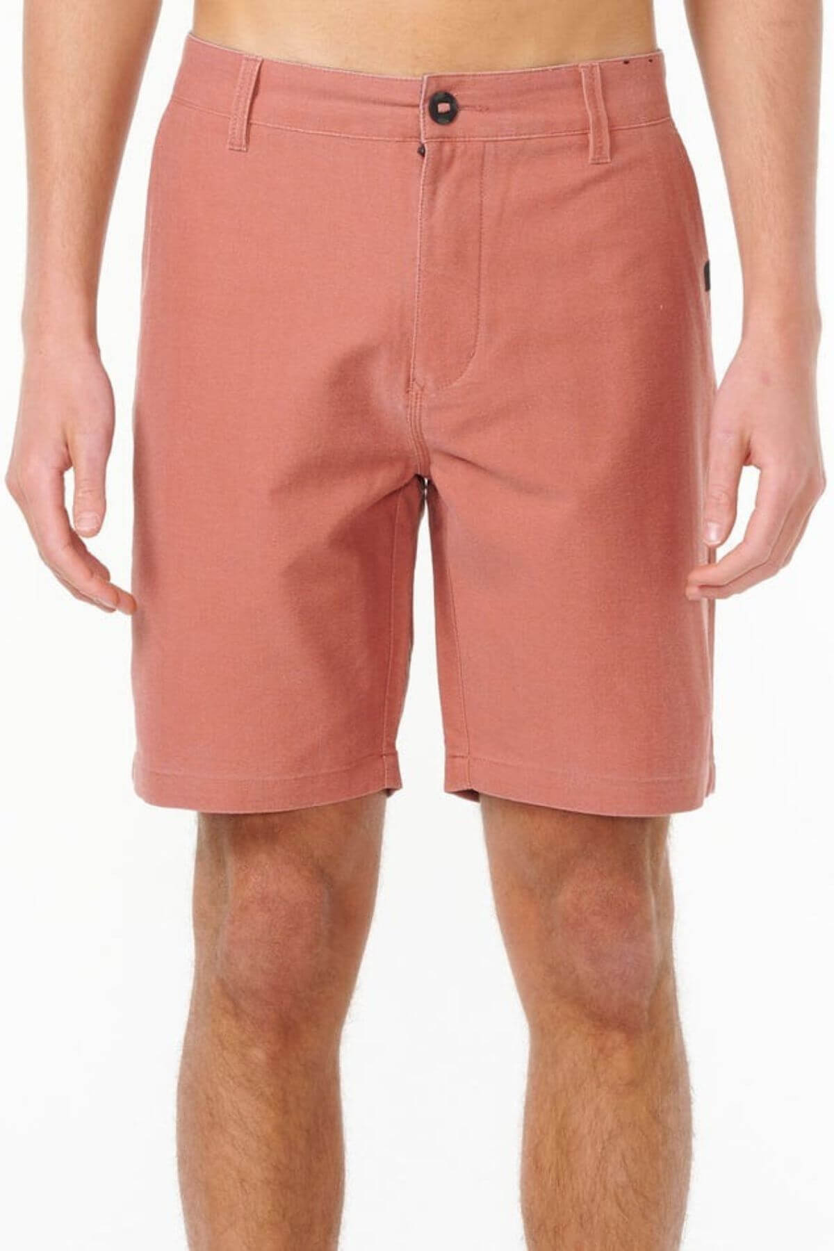 Someone wearing Rip Curl Boardwalk Epic Mix shorts in a light reddish-brown shade, with a vintage-inspired look and knee-length cut featuring a button and zipper closure, stands against a plain white background.