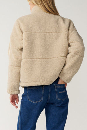 A person with long blonde hair is seen from the back against a plain light background, wearing blue jeans and a beige Rip Curl Cala Nights Sherpa Jacket featuring premium herringbone taping.
