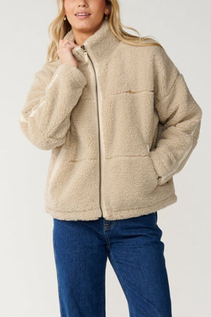 Someone wearing the Rip Curl Cala Nights Sherpa Jacket in light beige with a zip-up front and high collar, paired with dark blue jeans, stands against a plain background.