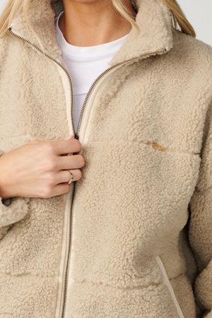 A person is shown from the shoulders down wearing the cream Rip Curl Cala Nights Sherpa Jacket over a white t-shirt, with one hand holding the zipper near the collar.