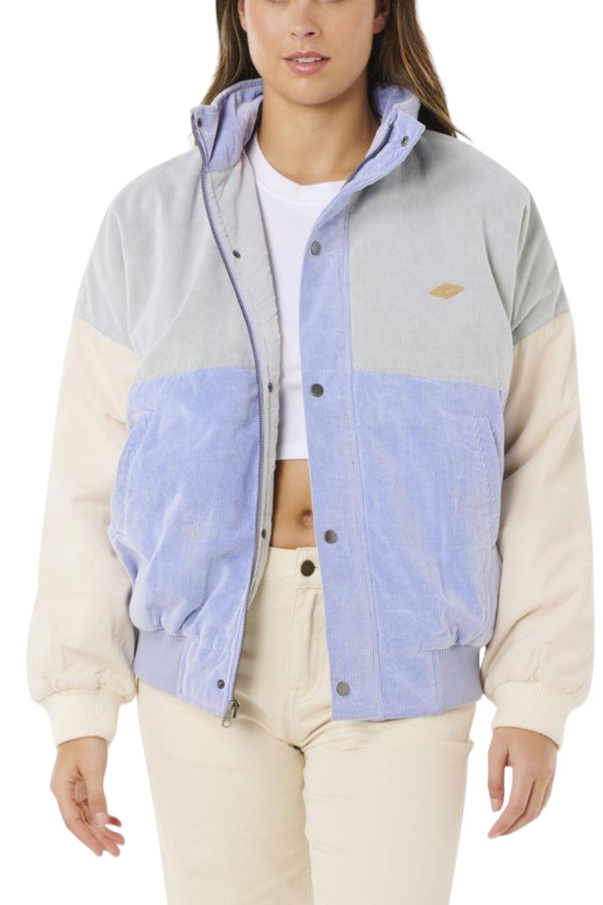 A woman wears the Rip Curl Cala Vadella Jacket, a retro style made from cotton corduroy in gray, light blue, and beige, layered over a white crop top and beige pants. The jacket is unzipped and her hands are at her sides.