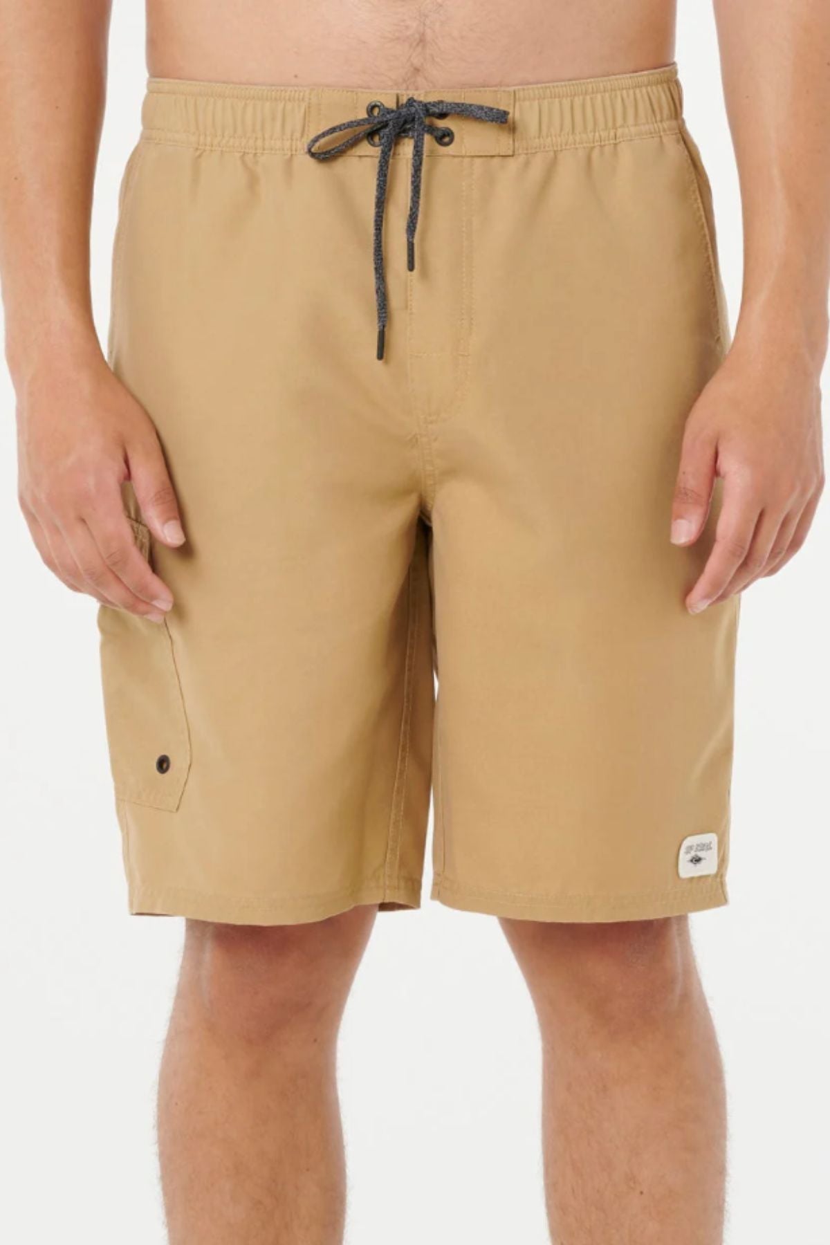 Someone stands against a white background wearing tan Rip Curl Classic Surf Cargo Shorts, featuring a black drawstring, side pocket, and a small white patch near the hem, resembling relaxed-fit cotton trail shorts.