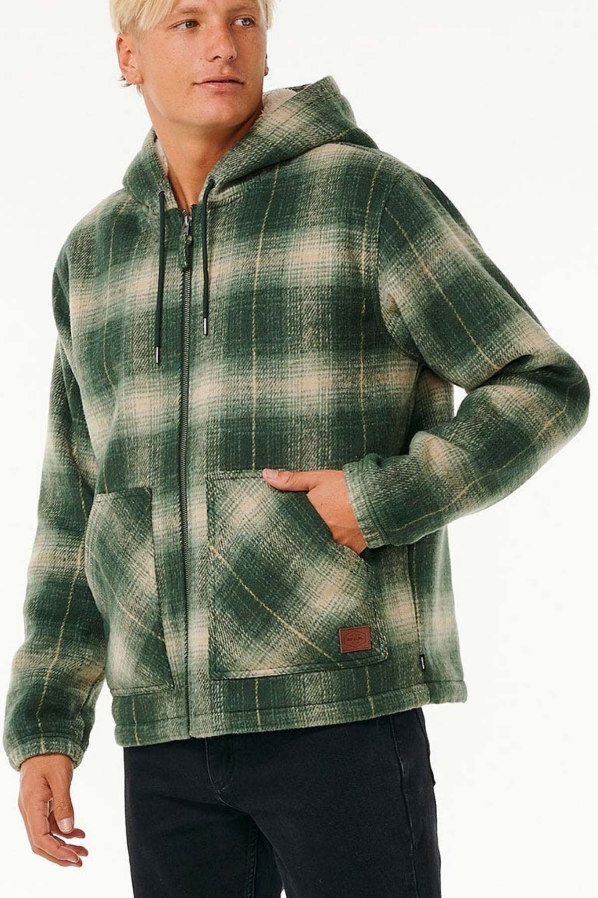 Wearing the Rip Curl Classic Surf Check Jacket in green and beige with front pockets and a sherpa-lined hood, a person stands with one hand in the pocket, looking to the side.