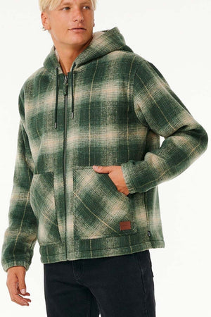 Wearing the Rip Curl Classic Surf Check Jacket in green and beige with front pockets and a sherpa-lined hood, a person stands with one hand in the pocket, looking to the side.