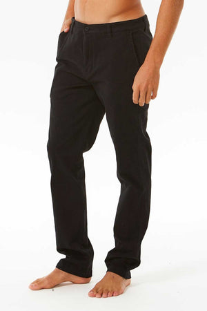 A person models Rip Curl Classic Surf Chino pants in black, shot waist down against a white background. Featuring a relaxed fit and stretch cotton twill with button closure, the barefoot individual has one hand in their pocket, capturing the essence of everyday wear.