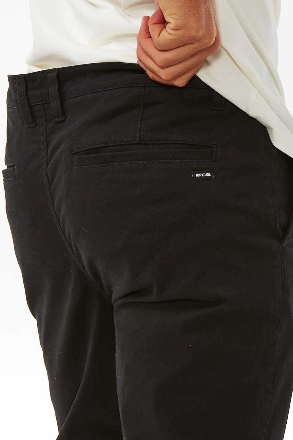 A person in black Rip Curl Classic Surf Chino pants and a white shirt, with one hand in the back waistband. The stretch cotton twill pants have a logo tag on the pocket.