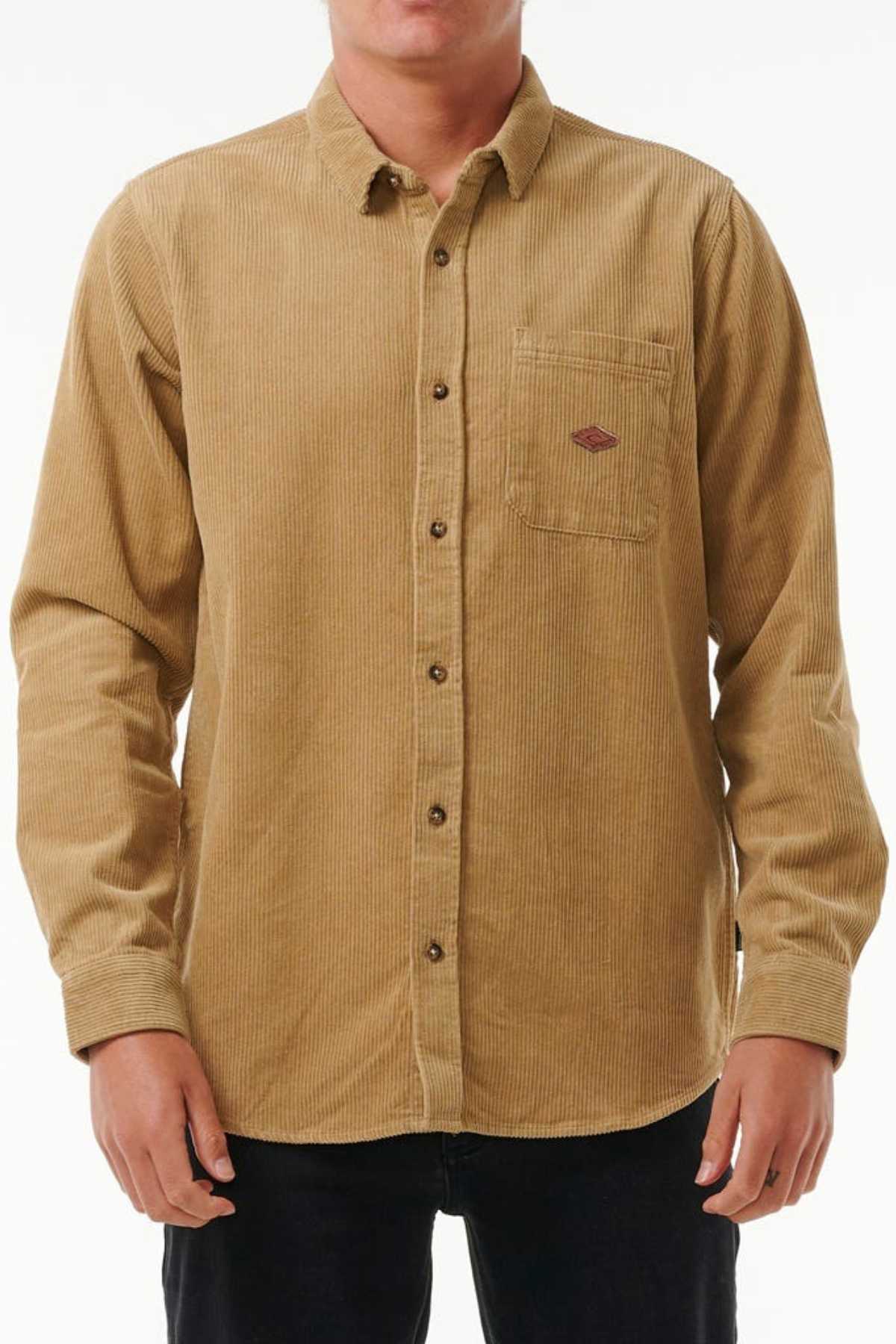 Sporting the Rip Curl Classic Surf Cord Shirt in tan corduroy with its button-up design, long sleeves, and a logo-adorned chest pocket, paired with black pants, the outfit pops against the white backdrop.