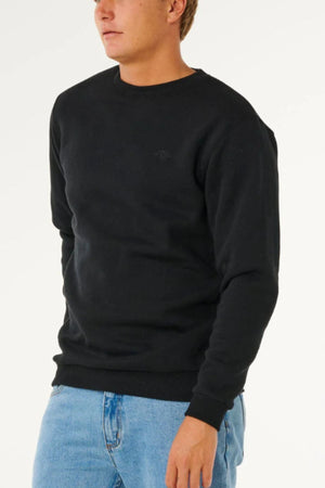 Wearing the Rip Curl Classic Surf Crew black sweatshirt with an embroidered logo and light blue jeans, a person stands against a white background, partially out of frame to highlight this surf essential style.