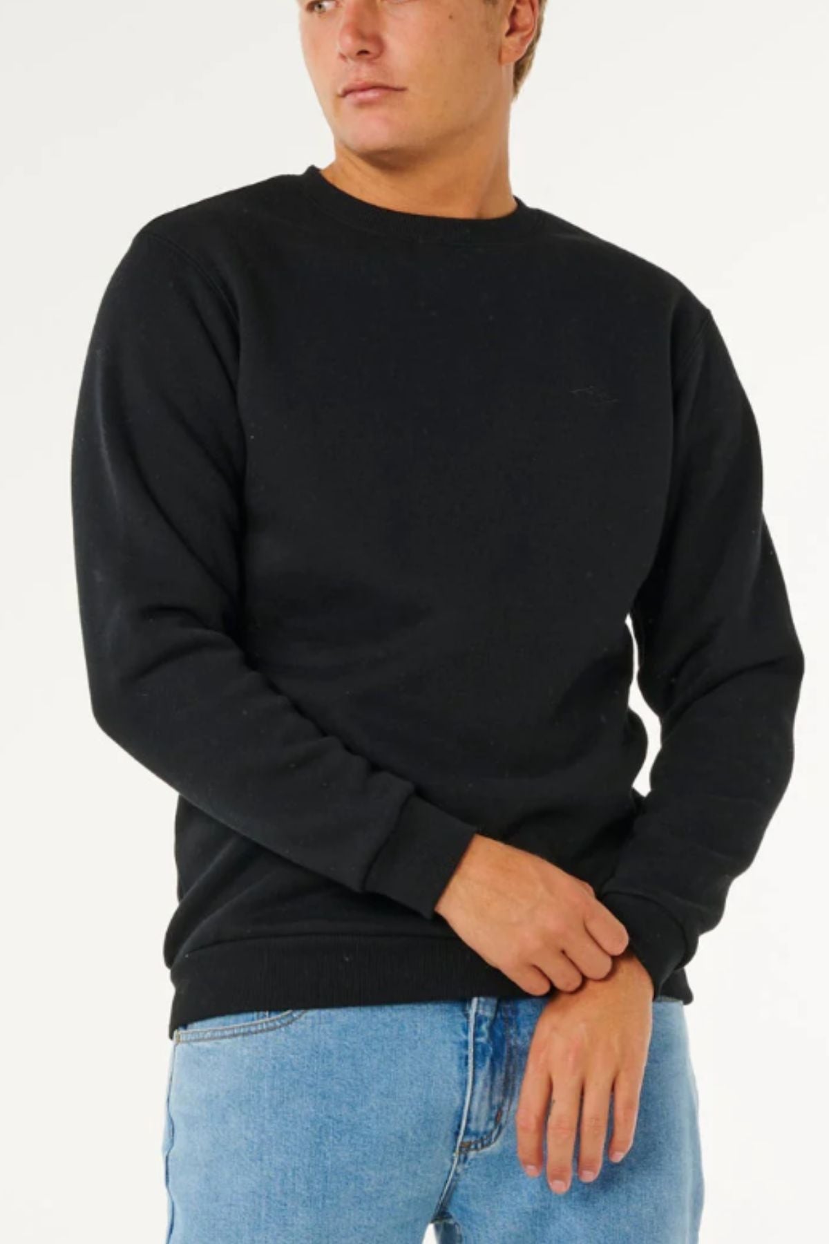A person wears a Rip Curl Classic Surf Crew black long-sleeve sweatshirt with an embroidered logo and light blue jeans against a white background—an essential surf-inspired addition to any wardrobe.