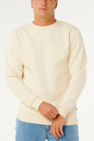 Wearing the Rip Curl Classic Surf Crew with an embroidered logo and light blue jeans, a person stands against a white background, hands clasped—an essential piece for surf style.
