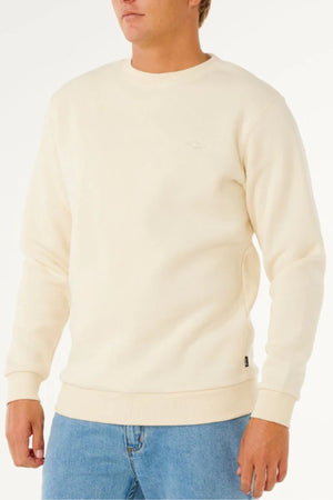 A person in a cream Rip Curl Classic Surf Crew sweatshirt with an embroidered logo and blue jeans stands against a white backdrop. Featuring a crew neck and ribbed cuffs, the surf essential is shown with the face partially out of frame.