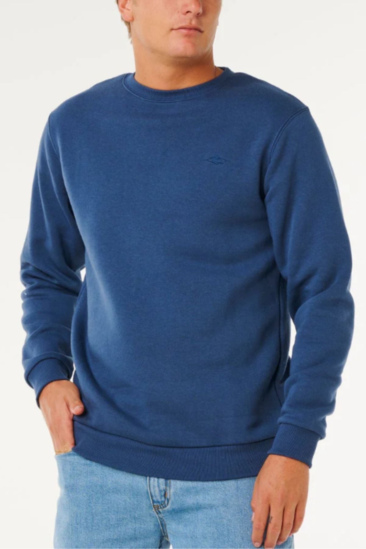 Wearing a Rip Curl Classic Surf Crew with an embroidered logo and light blue jeans, a person stands with one hand in their pocket against a white background—a true surf essential.
