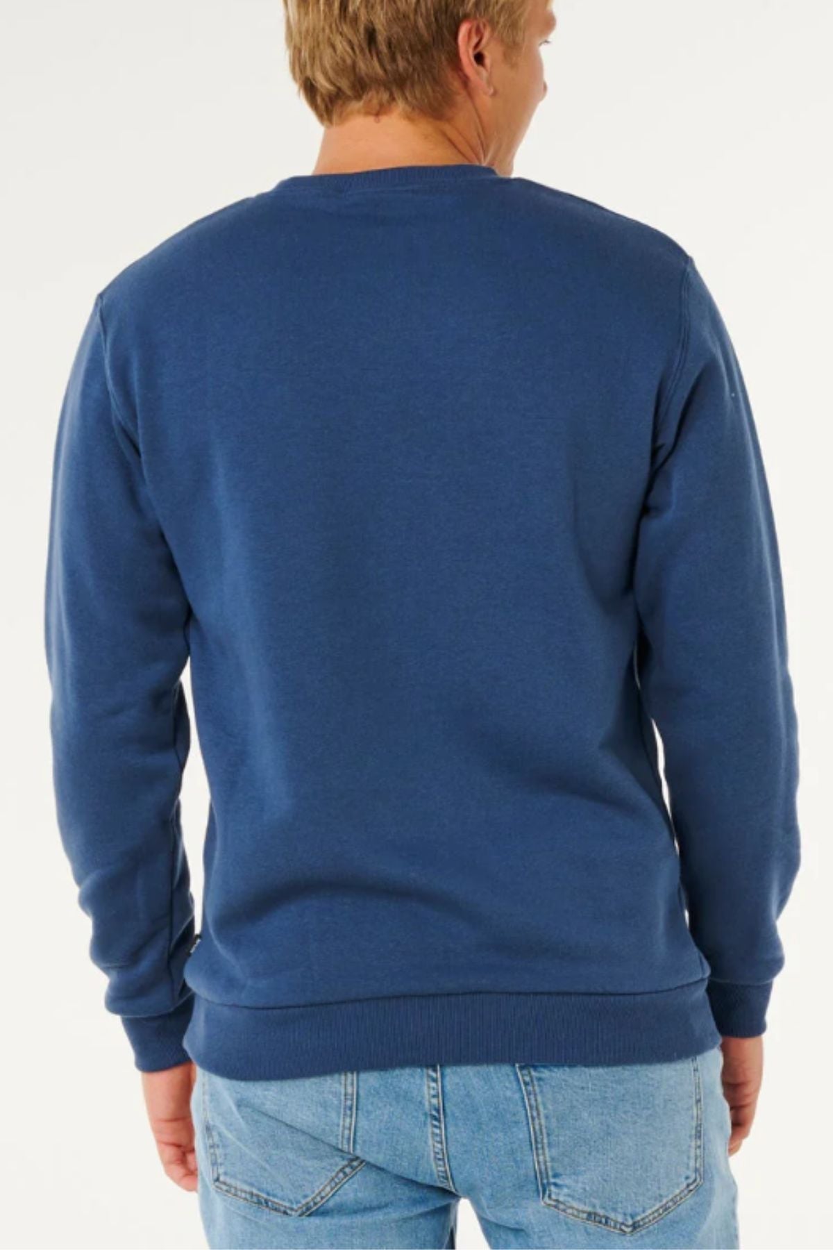 A person wearing a blue Rip Curl Classic Surf Crew sweatshirt with an embroidered logo and light blue jeans is shown from the back, standing against a plain white background.