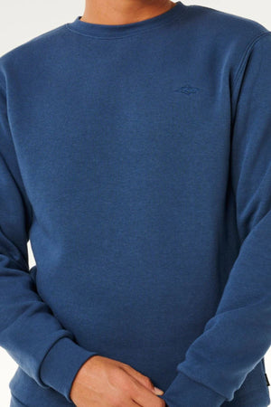 Wearing the blue Rip Curl Classic Surf Crew with an embroidered logo and long sleeves, a person stands with hands clasped in front against a plain, light background—capturing the true surf essential vibe.