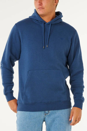 A person stands against a plain light background wearing a blue Rip Curl Classic Surf Hood with a front pocket and drawstrings, paired with light blue jeans.