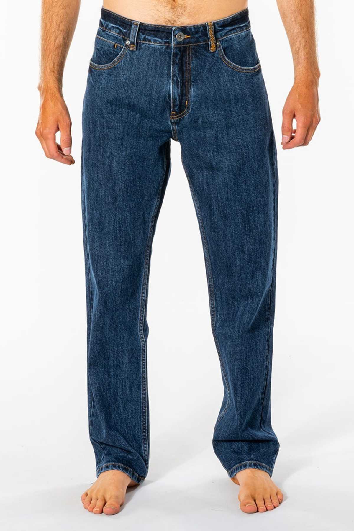 A person stands barefoot against a white background wearing Rip Curl Classic Surf Jeans. The focus is on the legs and lower torso, showing off the classic fit with stretch cotton denim, front pockets, and button closure.