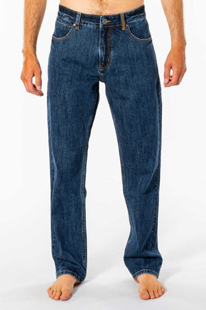 A person stands barefoot against a white background wearing Rip Curl Classic Surf Jeans. The focus is on the legs and lower torso, showing off the classic fit with stretch cotton denim, front pockets, and button closure.