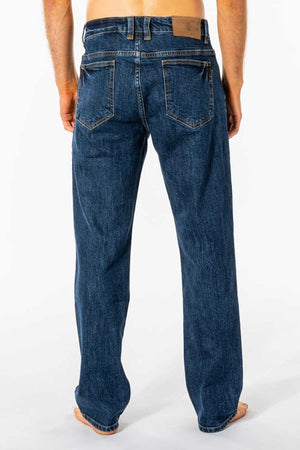 A barefoot person is seen from the back wearing blue Rip Curl Classic Surf Jeans, featuring a straight fit. These jeans have stretch cotton denim for comfort, two back pockets, visible stitching, and a brown leather patch on the waistband against a plain white background.