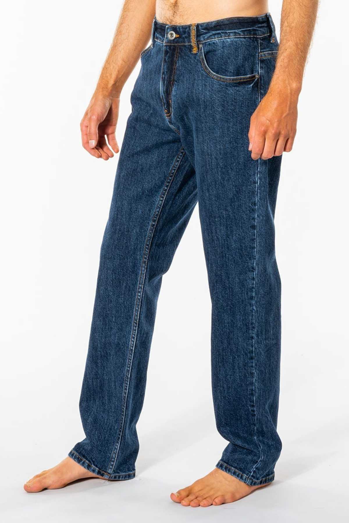 A person stands barefoot on a white background, wearing Rip Curl Classic Surf Jean. These blue jeans feature a classic straight fit made from stretch cotton denim for great comfort, with visible stitching and pockets. The upper body isn't fully visible.