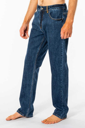 A person stands barefoot on a white background, wearing Rip Curl Classic Surf Jean. These blue jeans feature a classic straight fit made from stretch cotton denim for great comfort, with visible stitching and pockets. The upper body isn't fully visible.