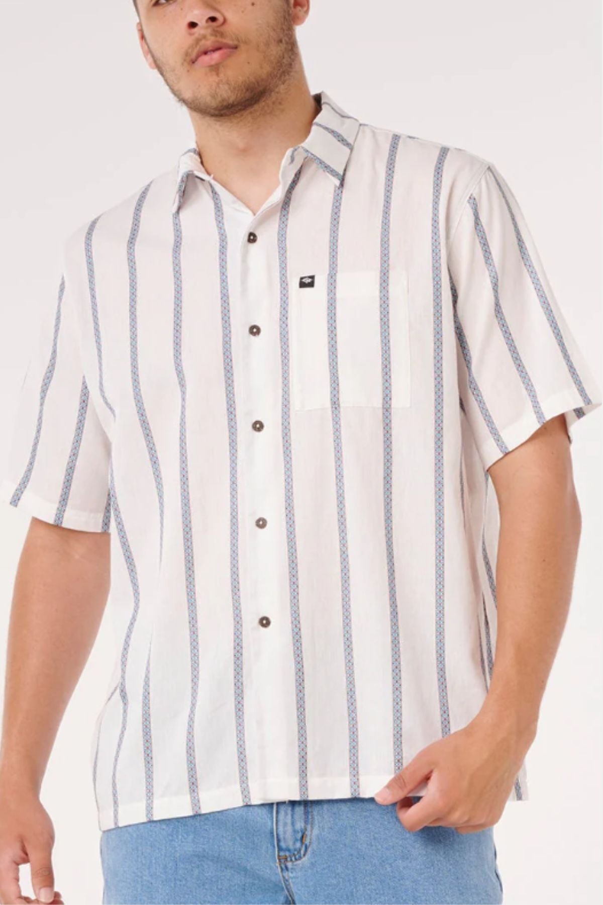 Wearing the Rip Curl Classic Surf Stripe Short Sleeve Shirt in white with blue and gray stripes and light blue jeans, a person stands against a plain light background.