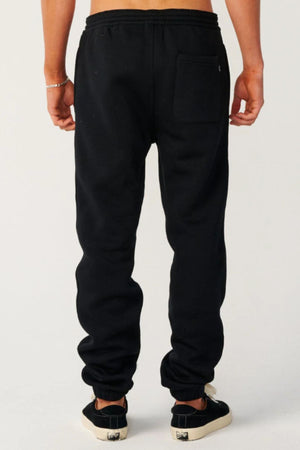 A person is seen from behind wearing Rip Curl Classic Surf Trackpant, black and white sneakers, and a silver bracelet on their left wrist. The plain light background emphasizes the relaxed style, ideal for casual wear.