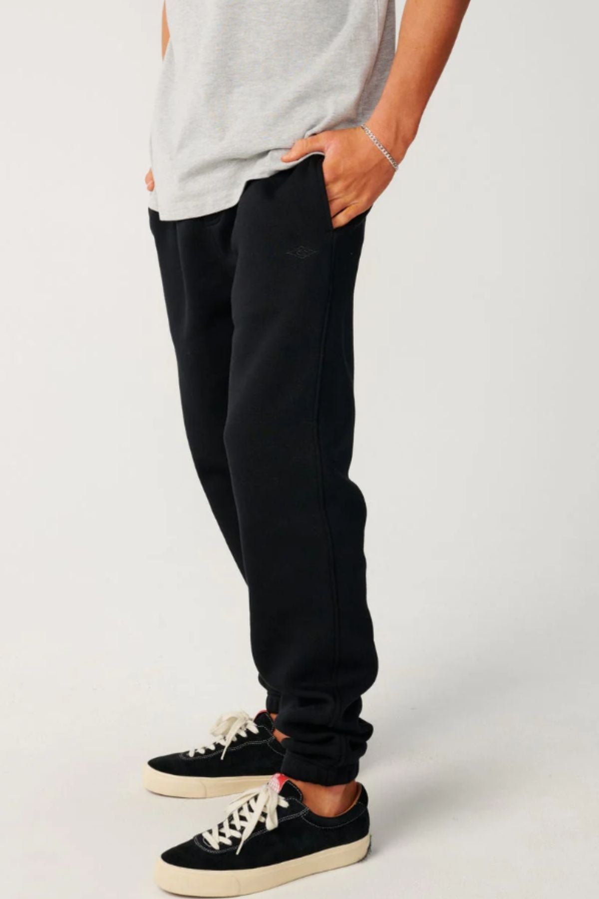 A person stands with hands in pockets, wearing a light grey t-shirt, Rip Curl Classic Surf Trackpant in black, and black low-top sneakers with white soles. The photo shows this casual look from shoulders to feet against a neutral background.