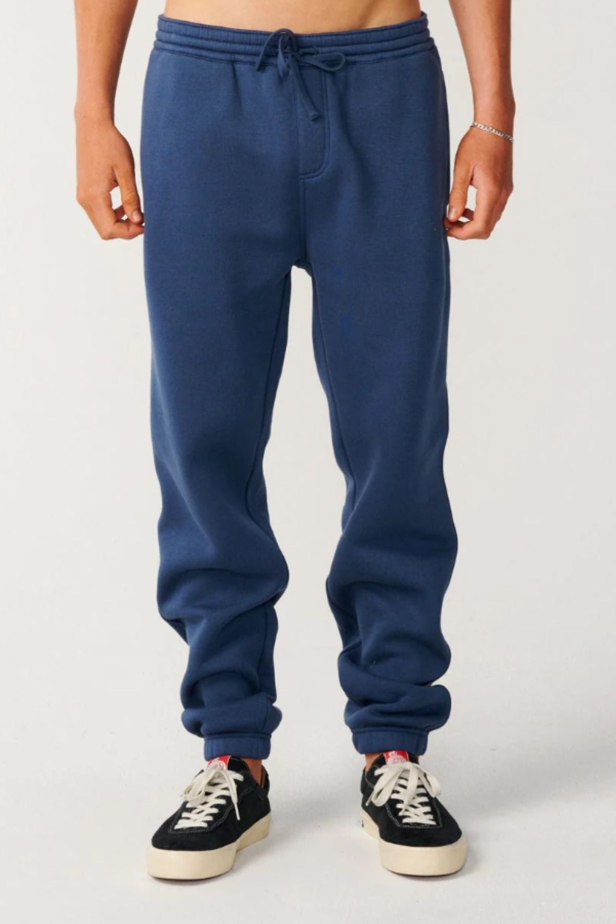 Wearing the Rip Curl Classic Surf Trackpant in blue with black-and-white sneakers, a person stands against a plain light background. Only the lower torso and legs are visible, drawing attention to this stylish casual wear.