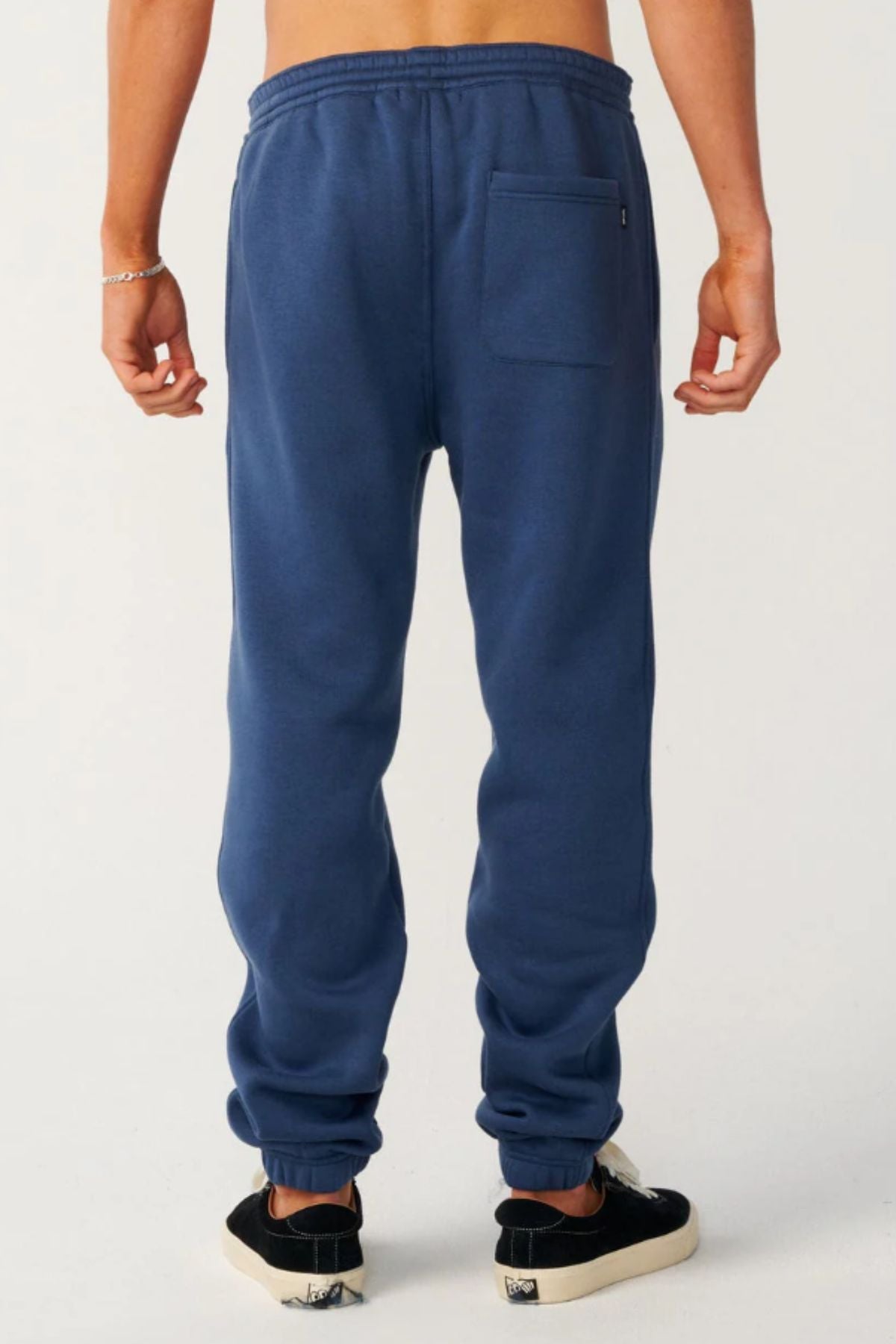 A person stands facing away, wearing Rip Curl Classic Surf Trackpant in blue with a back pocket—ideal for casual wear—paired with black slip-on sneakers and a silver bracelet, set against a plain light background.