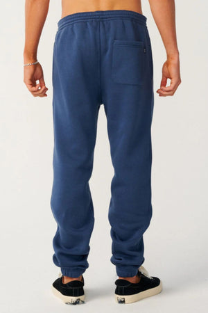 A person stands facing away, wearing Rip Curl Classic Surf Trackpant in blue with a back pocket—ideal for casual wear—paired with black slip-on sneakers and a silver bracelet, set against a plain light background.