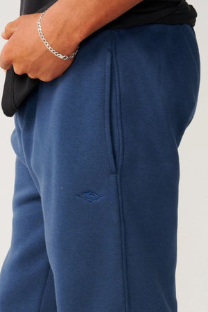 A person wears Rip Curl Classic Surf Trackpant in dark blue with a relaxed fit, right hand in the pocket showing a silver chain bracelet. The sweatpants feature a small embroidered logo near the pocket, ideal for casual wear.