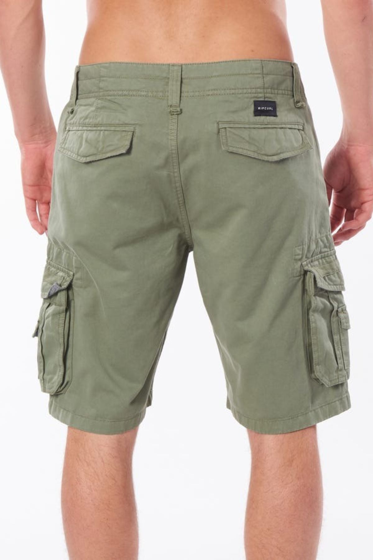 A person wearing Rip Curl Classic Surf Trail Cargo Short, crafted from 100% cotton, stands with their back to the camera. The olive green cargo shorts feature multiple buttoned pockets and a small black label on the right pocket, creating a unique contrast against the plain white background.