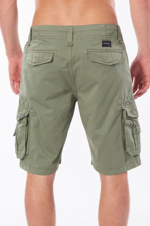 A person wearing Rip Curl Classic Surf Trail Cargo Short, crafted from 100% cotton, stands with their back to the camera. The olive green cargo shorts feature multiple buttoned pockets and a small black label on the right pocket, creating a unique contrast against the plain white background.