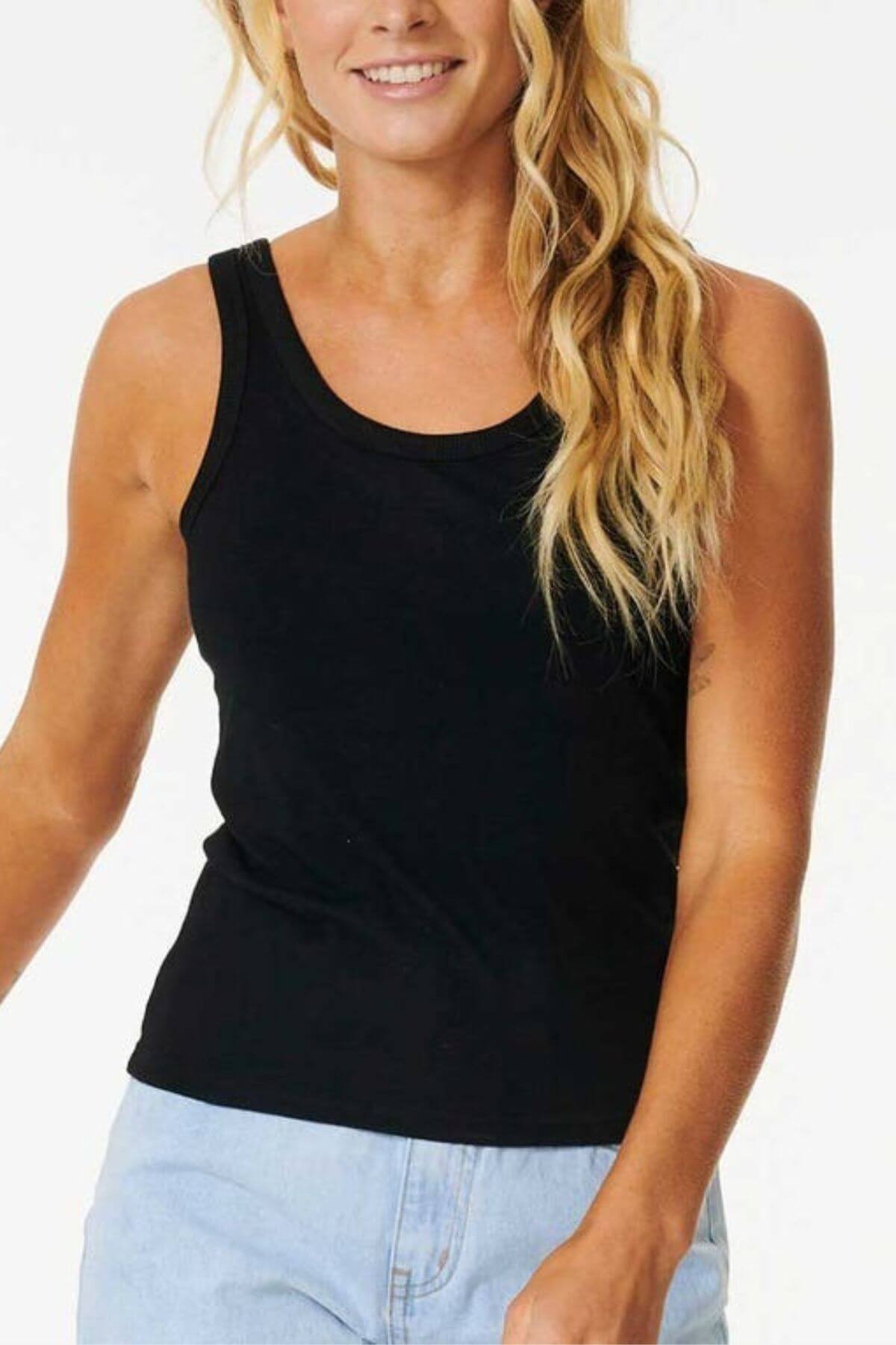 A woman with long, wavy blonde hair is smiling while wearing a sleeveless Rip Curl Classic Tank and light blue jeans. She stands against a plain white background, with only her upper body visible.