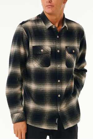 Someone stands against a plain white background wearing the Rip Curl Count Flannel Shirt, a black and beige plaid long-sleeve cotton flannel featuring two chest pockets.