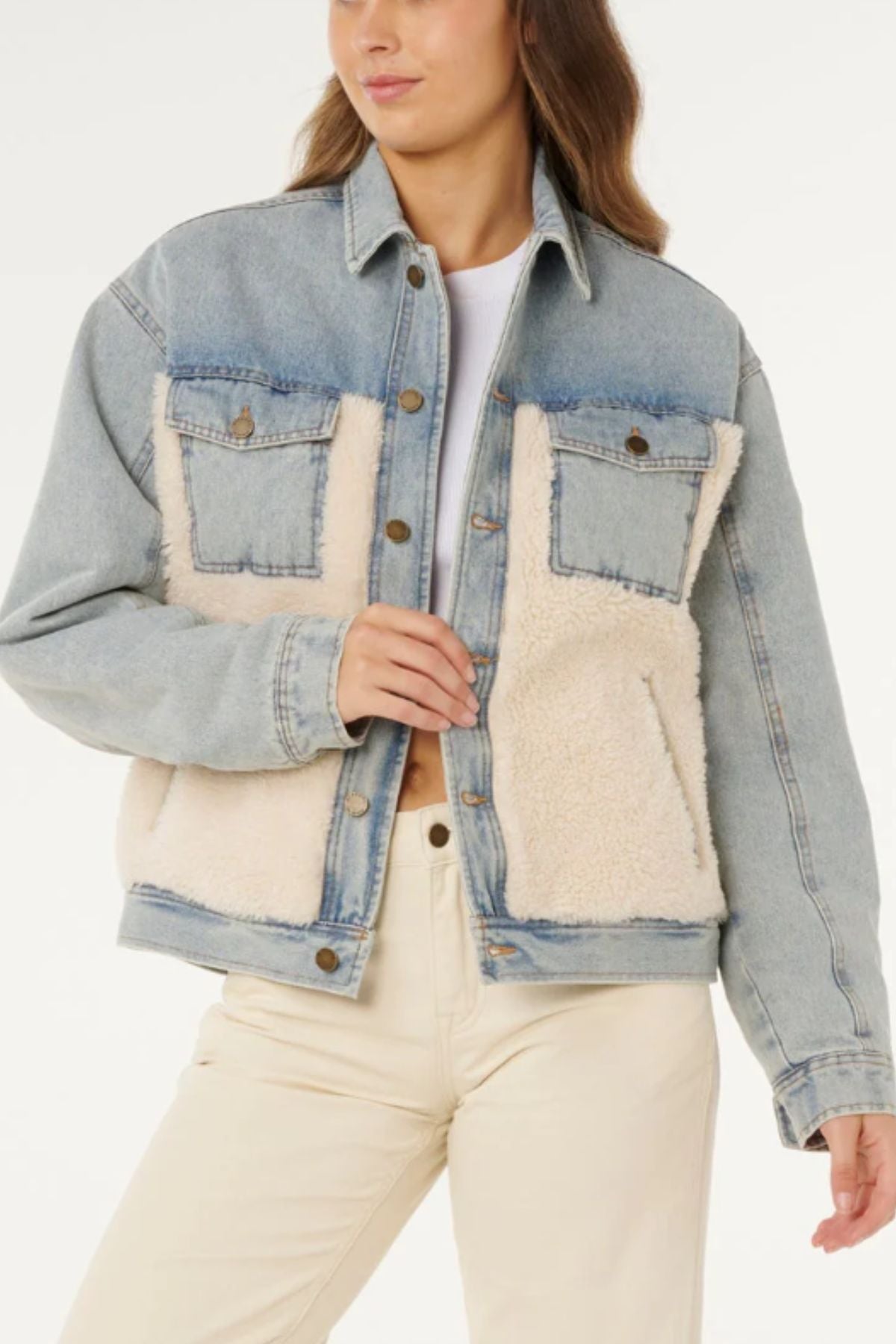 A woman wears the Rip Curl Cruisin Sherpa Denim Jacket in light blue, featuring cream sherpa panels, quilted lining, metal buttons, chest pockets, and a relaxed fit, styled with off-white pants and a white top.