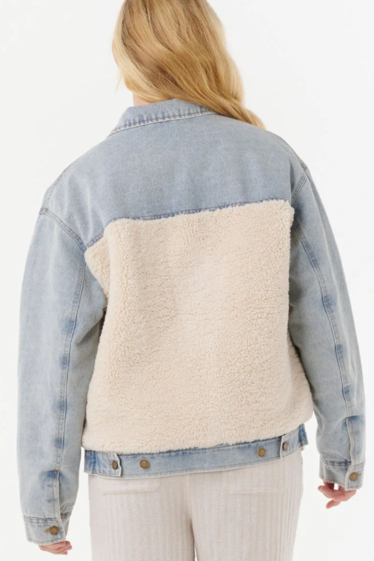 Someone with long blonde hair models the Rip Curl Cruisin Sherpa Denim Jacket in light blue, showcasing its large cream sherpa panel and quilted lining, paired with light pants, seen from behind—ideal for a laid-back weekend look.