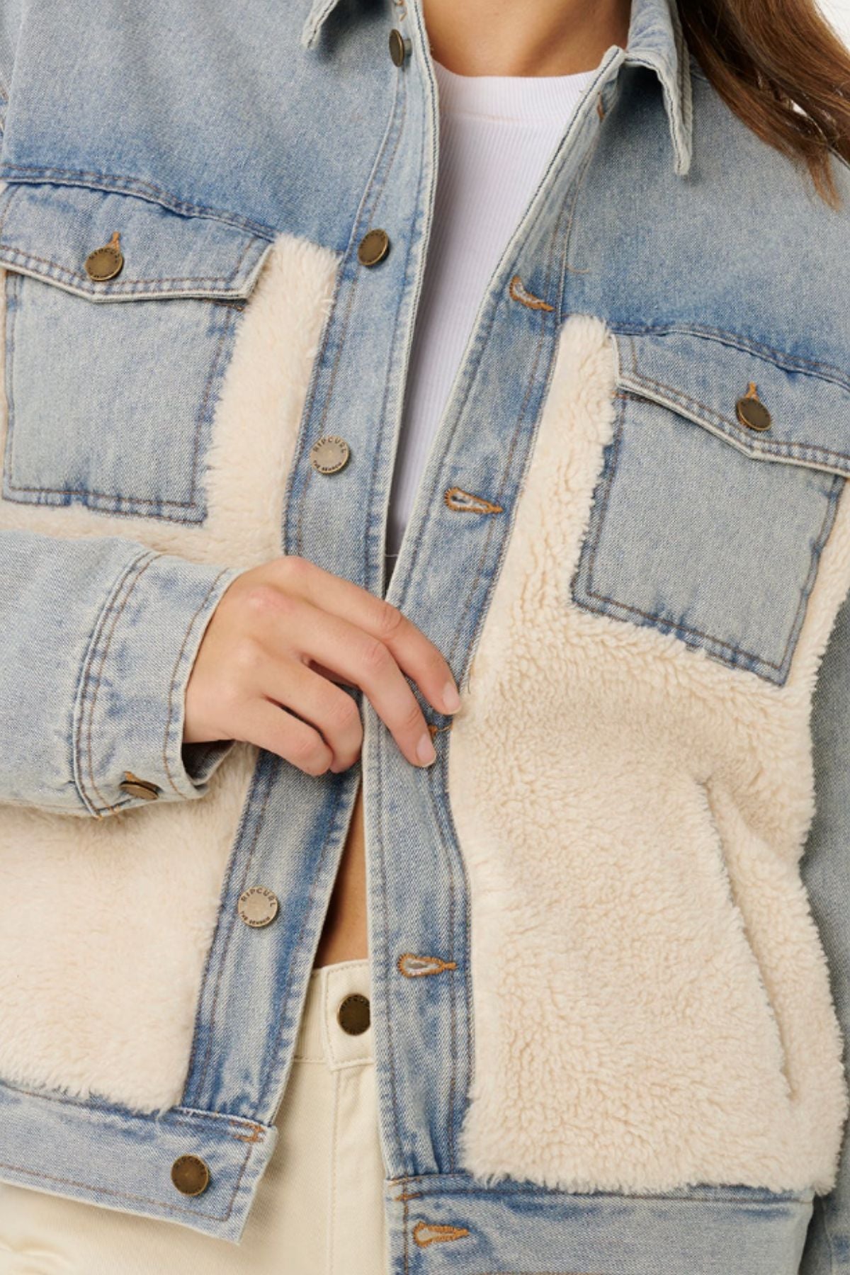 A person wearing the Rip Curl Cruisin Sherpa Denim Jacket, featuring beige sherpa panels, brass buttons, chest pockets, and quilted lining over a white top. Only the torso and one hand adjusting the jacket are shown—ideal for casual weekends.