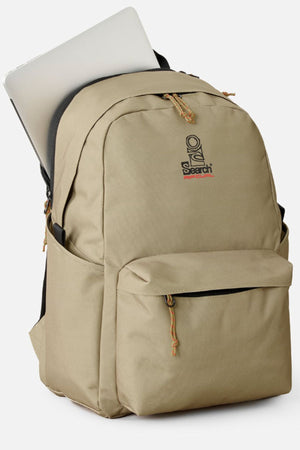 The Rip Curl Crusha 30L Search Backpack in tan, featuring the "Search" logo, has a front zippered pocket, side pocket, and holds a silver laptop partially visible in the main compartment.