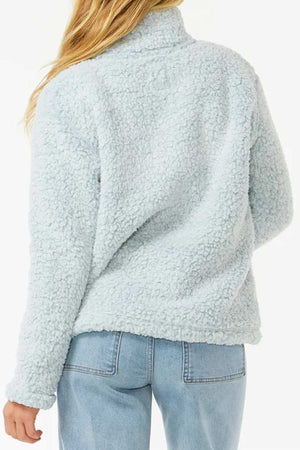A person with long blonde hair is shown from the back, wearing a light blue Rip Curl Dark N Stormy 1/4 Zip Crew fleece sherpa jacket and light blue jeans. The background is plain white.