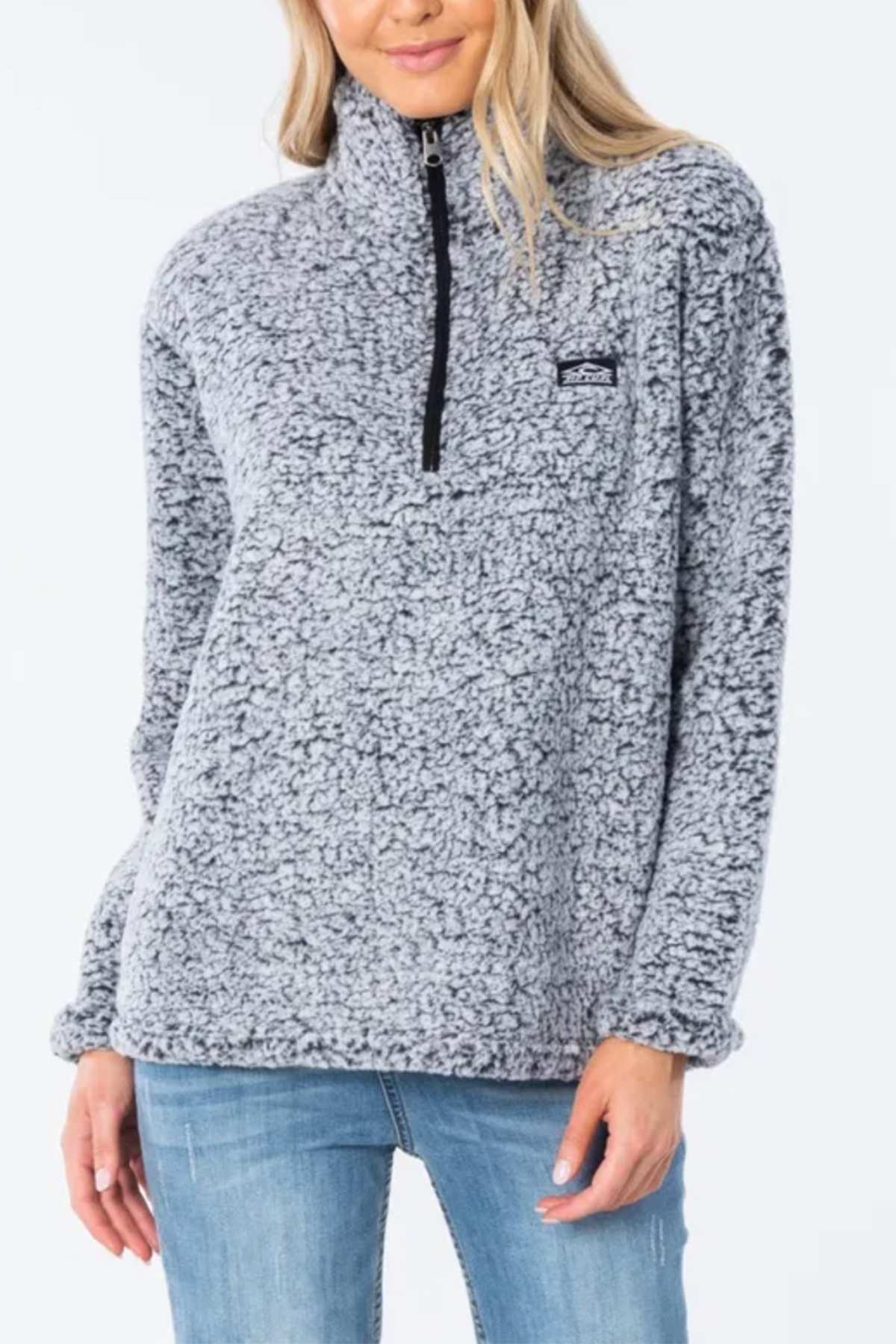 A woman stands against a plain light background in blue jeans and a light gray Rip Curl Dark N Stormy 1/4 Zip Crew made from cozy poly marble fleece with a quarter-zip neckline.