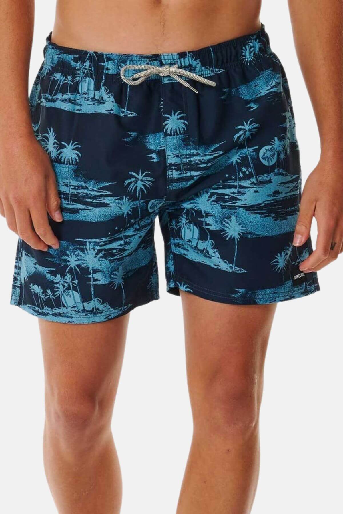 A person wears the Rip Curl Dreamers Volley—navy blue board shorts with a light blue palm tree print, white drawstring, made from recycled rip suede—shown waist to mid-thigh against a plain background.