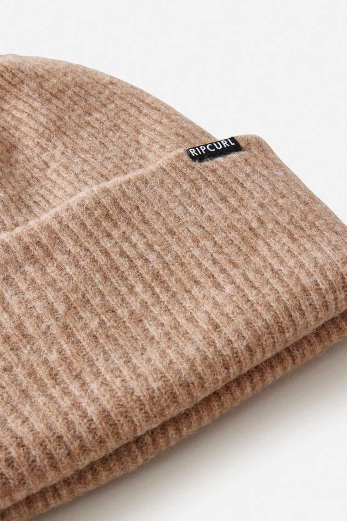 A close-up of the Rip Curl Eclipse Tall Beanie in light brown, made from soft acrylic yarn with a small black "Rip Curl" label on the folded edge. Laid flat on a white surface, it's perfect for adding warmth and style to your winter look.