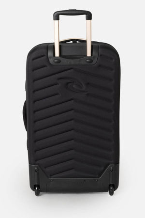 The Rip Curl F-Light Global 100L Soleil Travel Bag in black has a zigzag textured front, is made from recycled polyester, features an extended telescopic handle, two wheels for easy maneuverability, and internal compression straps. It stands upright against a plain gray background.
