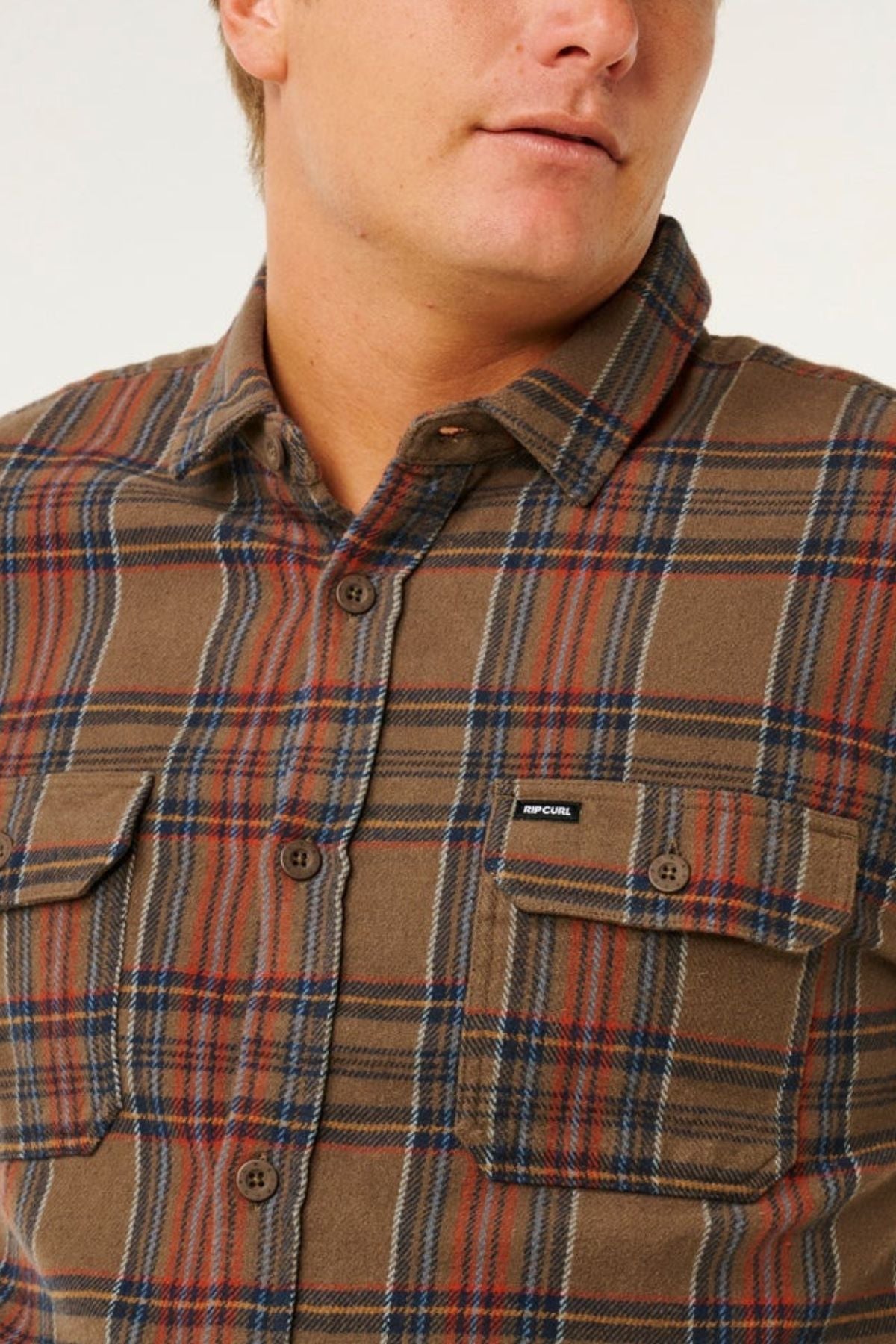 A person wears the Rip Curl Griffin Flannel Shirt, a men’s brown and red plaid cotton flannel with chest pockets and a small Rip Curl label, shown cropped at the upper torso and part of the face.
