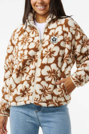 A person is wearing the Rip Curl Hibiscus Heat Polar Fleece Zip Through jacket in brown and white floral, paired with light blue jeans. The jacket has a black and white chest patch, and they smile with their hands in the pockets.