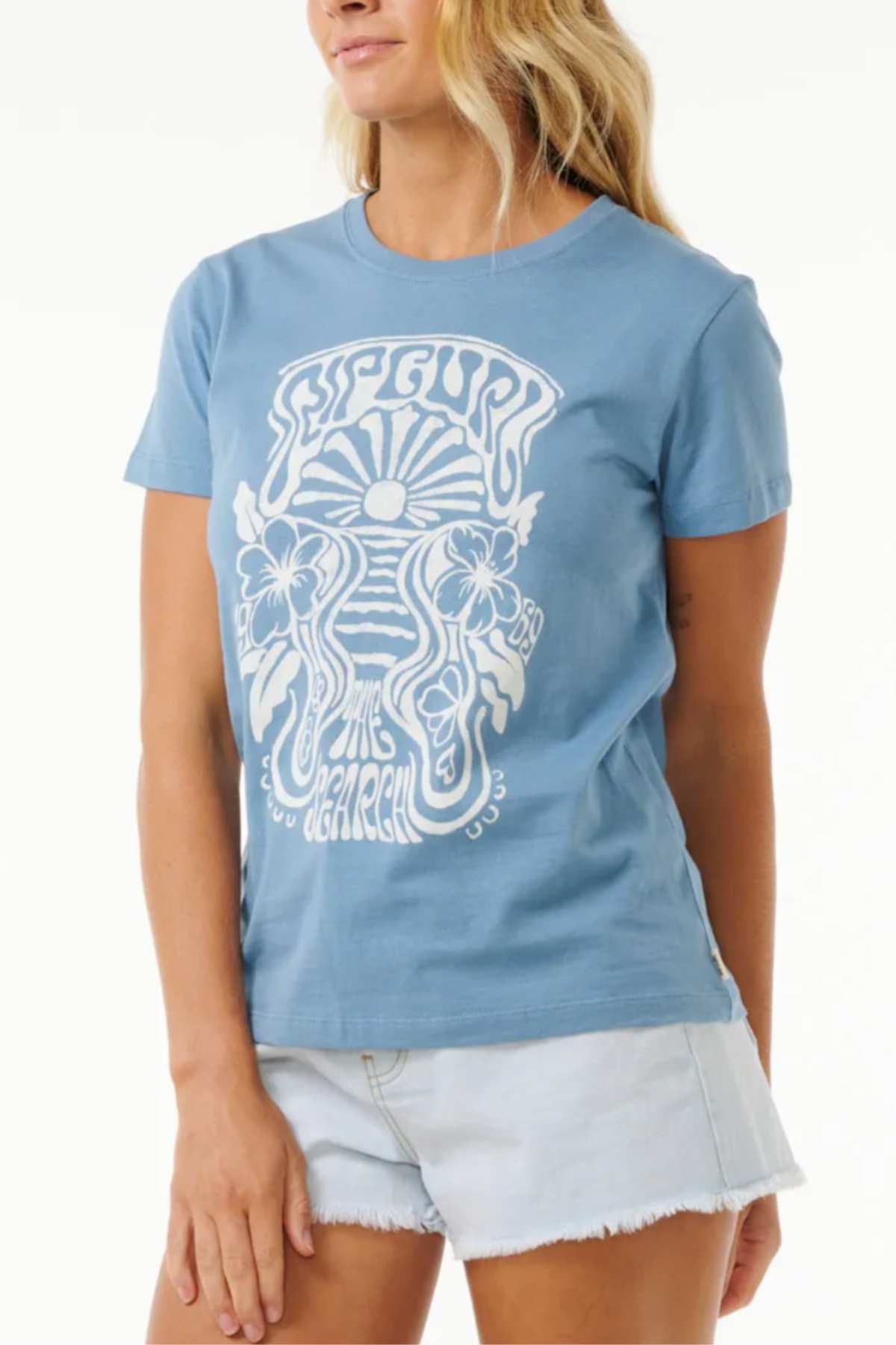 A person wearing the Rip Curl High Tide Psychic Standard Tee, a light blue graphic T-shirt made from soft cotton jersey fabric with a white, intricate design featuring the words "RIP CURL High Tide," also dons light denim shorts. They have long, wavy blonde hair and stand against a plain white background.