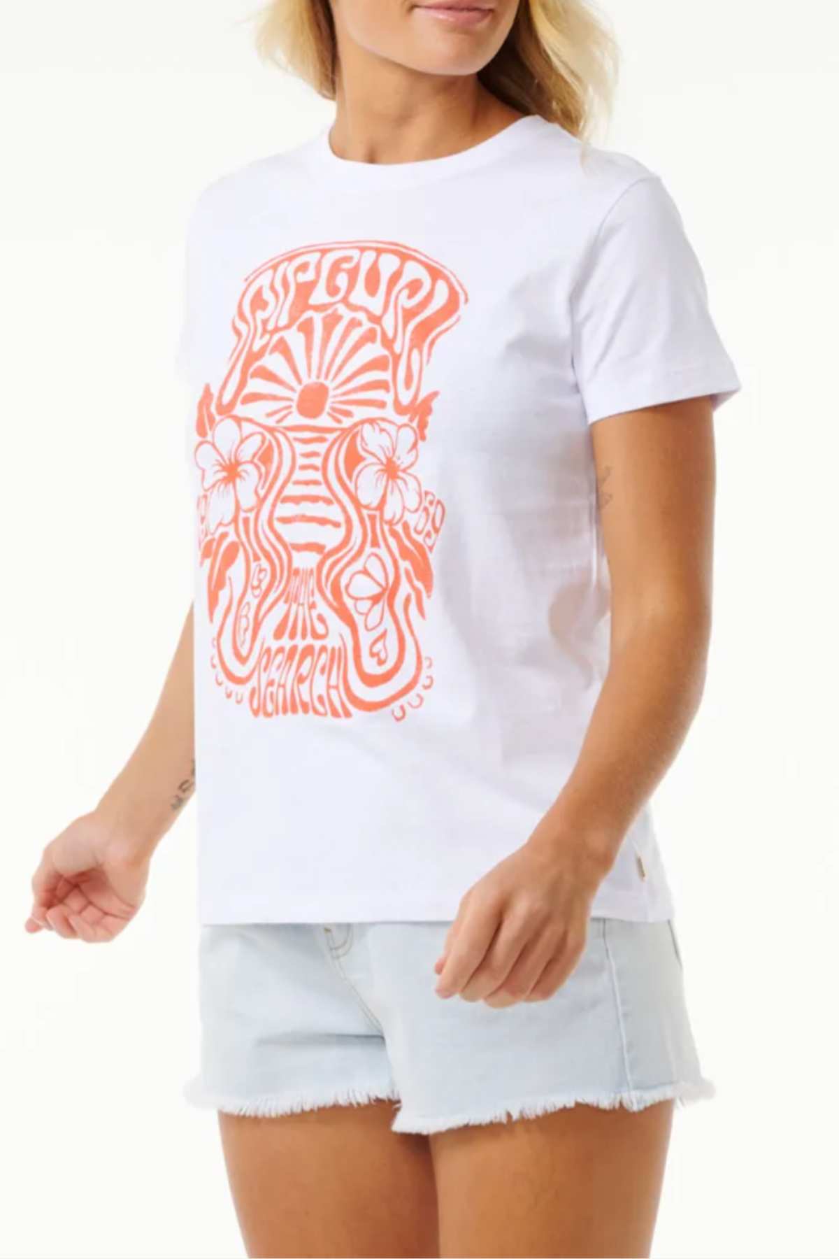 A person wearing the Rip Curl High Tide Psychic Standard Tee, a white t-shirt crafted from soft cotton jersey fabric, showcases an orange psychedelic-style graphic design with "Rip Curl High Tide" and floral elements. Paired with light blue denim shorts, the image focuses on their upper body and midsection.